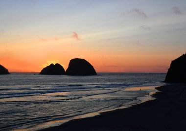 Oceanside Beach, Oregon 'daki plaja gidenler, parlayan turuncu günbatımının tadını çıkarın. Deniz yığınları siluetlidir.