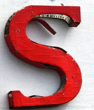 Büyük, kırmızı ahşap S beyaz reklam panosuna asılır. Boya yıpranmış ve soyulmuş..