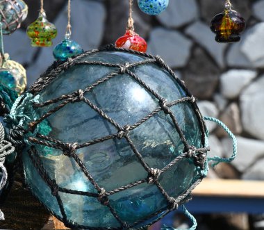 Büyük bir el patlaması, cam şamandıra dışarıda güneşin altında duruyor. Cam su renginde ve düğümlü ipe sarılı..