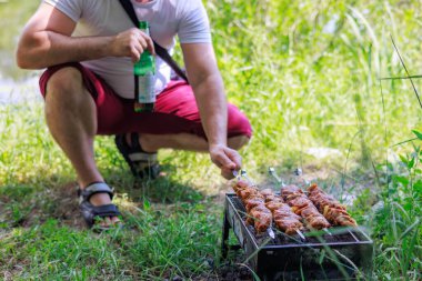 Elinde bira şişesi olan bir adam et kızartıyor.