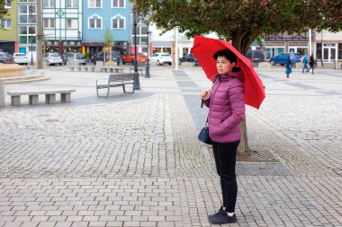 Kırmızı şemsiyeli bir kadın çiçek açan bir ağacın yanındaki meydanda duruyor..