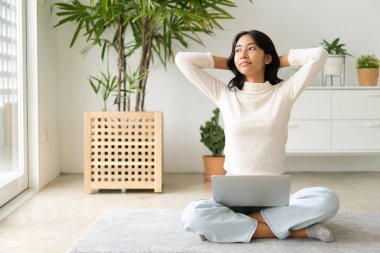 Asian woman relax  with laptop hands behind head  while sitting on the floor in morning sunlight at home