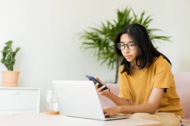 Asian woman wear glasses typing work on laptop. Asia student learn via internet with computer, using mobile phone,  while sitting on sofa in living room at home