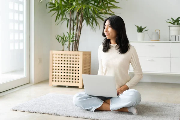 Asian woman relax from using laptop and looking out window. Asia student typing computer while sitting on the floor in morning at home