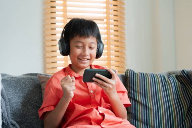 Excited Asian boy playing online game on smart phone with headphones. Asia man using mobile phone happy smile at home  in morning 