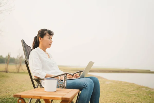 Asian woman typing on laptop while sitting at campsite outdoor. Asia women relax working using computer amidst meadow field and river.  Concept camping of lifestyle vacation and freedom