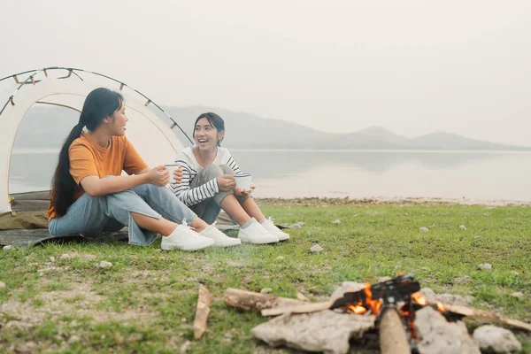 亚洲两名少女坐在草地上聊天 夕阳西下时举着玻璃窗搭帐篷 快乐的亚洲少女在户外露营地享受 免版税图库图片