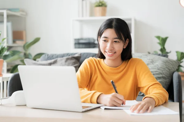 在家学习 亚洲女孩在网上通过网络学习视频会议 电脑辅导 亚洲女人在家里早上使用笔记本电脑 在客厅做作业 图库照片