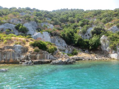 Rocky Akdeniz manzarası. Yoğun doğal bitki örtüsü ve kaya oluşumları sahile iniyor. Dağlar, çalı ve yeşilliklerle kaplı. Resmedilmeye değer ve vahşi bir görüntü sergiliyorlar.