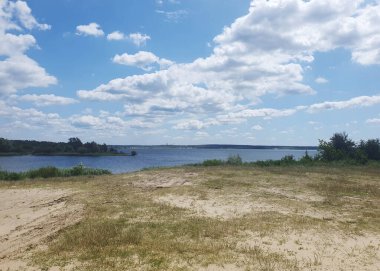 Açık mavi göl, sazlıklar ve kumlu kıyılar açık bir yaz gökyüzü altında yeşil ormanın arka planına karşı. Barışı, saflığı ve doğanın güzelliğini iletir. Açık hava eğlencesi, ekoloji, turizm temaları için