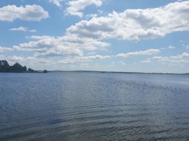 Açık mavi göl, sazlıklar ve kumlu kıyılar açık bir yaz gökyüzü altında yeşil ormanın arka planına karşı. Barışı, saflığı ve doğanın güzelliğini iletir. Açık hava eğlencesi, ekoloji, turizm temaları için