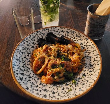 Dekoratif tabakta servis edilen iştah açıcı deniz ürünleri makarnası. Ayrıca masada açık bir mojito bardağı, bir bardak ve ahşap çubuklu bir kap var. Lokanta yemeği için rahat bir ortam yaratır.