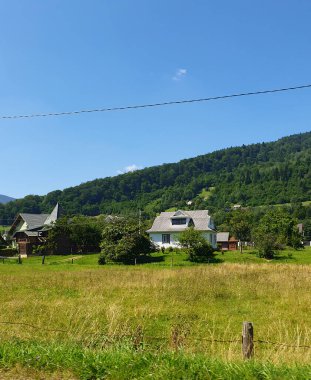 Güneşli bir yaz gününde Karpatlar 'ın rahat köyü. Yeşil çayırlar arasında geleneksel evler ve orman dağlarının arka planına karşı bahçeler. Parlak mavi gökyüzü, saf doğa bir barış ortamı yaratır.