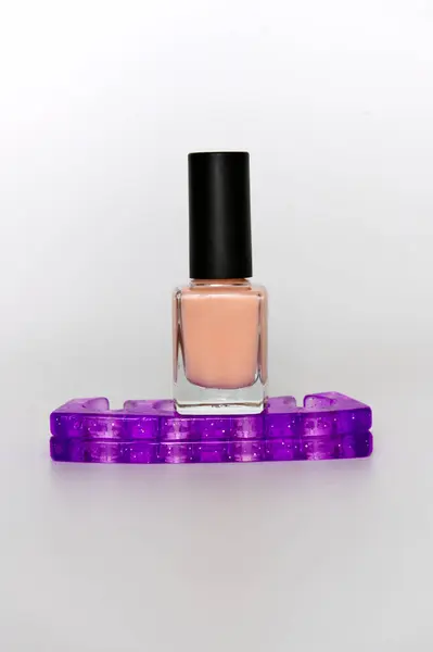 Bir şişe şeftali tırnak cilası beyaz zemin üzerinde sarmal mor bir zincir ile çevrilidir. Renklerin zıtlığını ve canlılığını vurgular.