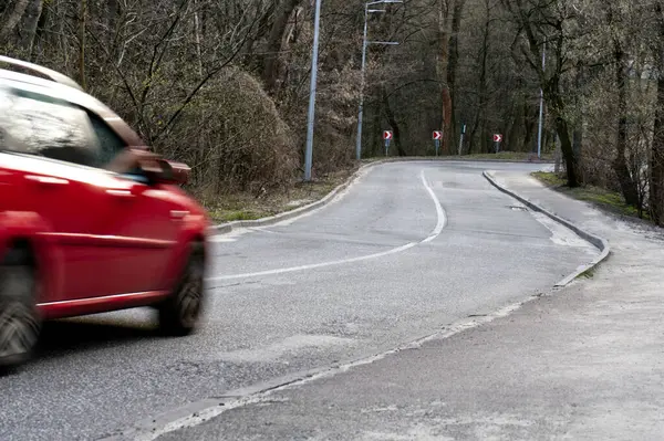 Parlak kırmızı bir araba, yemyeşil ve çiçek açan çiçeklerle çevrili, yılanımsı bir dağ yolunda hızla ilerliyor. Günbatımının renkleriyle boyanmış gökyüzü, bulutlu atmosfere hayat veriyor..