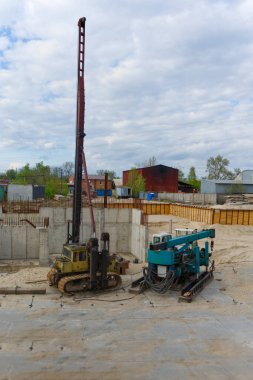 Görüntü, bulutlu bir gökyüzünün altında kazık çakan bir sürücü ve yardımcı ekipman bulunan bir inşaat alanını gösteriyor. Bu da, muhtemelen temel atmak için devam eden ağır inşaat çalışmalarını gösteriyor..