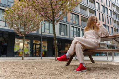 Bej kıyafetli bir insan, şehir ortamının ortasında ahşap bir bankta oturur. Çiçek açan pembe ağaçlarla modern mimariye biraz doğa katar.