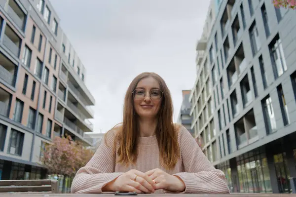 Resim masada oturan bir kişiyi tasvir ediyor. Modern binalar arka planı oluşturur, kentsel bir ortam önerir..