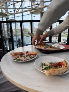 Bir insan, parlak, havadar bir kubbenin içindeki mermer bir masanın üzerindeki beyaz bir tabaktan bir dilim pizzaya uzanır. Sahne, doğal ışığın içeri aktığı sıradan bir yemek anını yakalıyor..