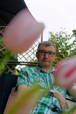 Adam ön plandaki bulanık pembe çiçeklerle kısmen gizlenmiş bir şekilde dışarıda oturuyor. Yeşil beyaz bir gömlek ve sol bileğinde bir saat giyiyor..