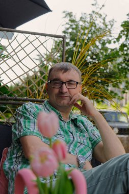 Bir insan, yemyeşil bitkiler ve canlı pembe lalelerle çevrili bir teras sandalyesinde rahatlar. Huzur ortamında sükunet ve mahremiyet vurgulanıyor.