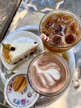 Serinletici buzlu bir kahve, karışık latte sanatıyla sıcak bir kahve, bir dilim katmanlı kek ve masadaki küçük bir pasta, rahatlatıcı bir ikindi ikramı için mükemmel..