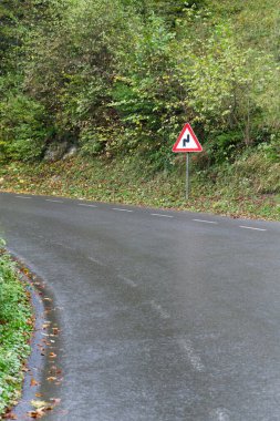  Görüntü, önünde keskin virajlar olduğunu gösteren bir uyarı işareti olan ıslak bir yol gösteriyor. Yol sola doğru kıvrımlı, ve tabela sağ tarafta, yoğun yeşilliklerle çevrili..