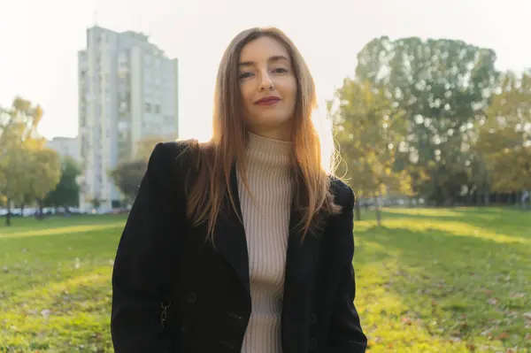 Bir kadın parkta dikiliyor, beyaz balıkçı yaka bir kazağın üzerine siyah bir ceket giyiyor.