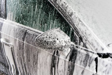 Bir araba yıkama yeri, bir aracın yan aynasını ve sürücü kapısının kalın sabun köpüğüyle kaplı olduğunu gösteriyor..