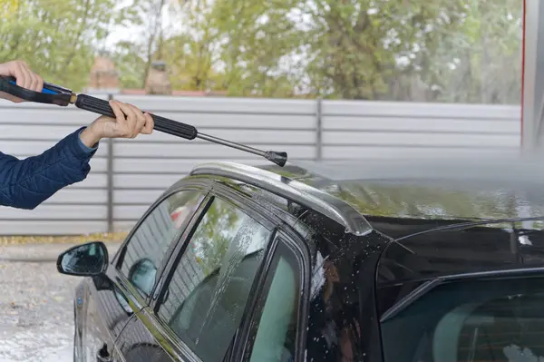 Görüntü, sabun ve suyla kaplı siyah bir arabayı temizlemek için yüksek basınçlı su hortumu kullanan birini gösteriyor. Görüntü, sabun köpüğüyle kaplı bir arabanın arka tarafını çekiyor, muhtemelen araba yıkama sırasında.. 