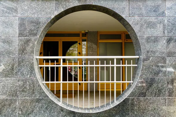 Görüntü, taş bir duvarda, ahşap kapılı ve pencereli bir binanın girişini gösteren dairesel bir açıklık gösteriyor. Bir metal korkuluk açığı geçer, geometrik kontrastı ekler.