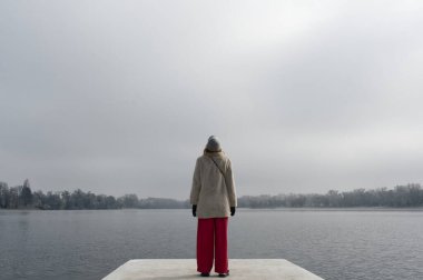 Bir insan bulutlu bir gökyüzünün altında sakin bir göle bakan beton bir platformda duruyor..