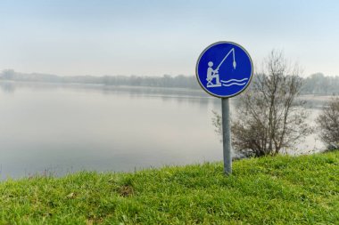 The image shows a sign indicating a designated fishing area by a calm lake. The serene surroundings and clear waters make it an inviting spot for fishing enthusiasts to relax and enjoy the activity. clipart