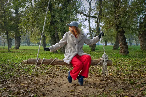 Bir kadın, ağaçlar ve düşen yapraklarla çevrili bir parktaki ahşap salıncakta huzurlu bir anın tadını çıkarıyor..
