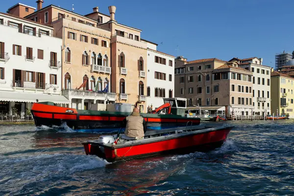 Venedik 'te renkli binalar ve teknelerin yer aldığı sakin bir kanal. Küçük kırmızı bir teknede bir kişi ve arka planda daha büyük bir kırmızı tekne resim gibi bir sahne oluşturur..