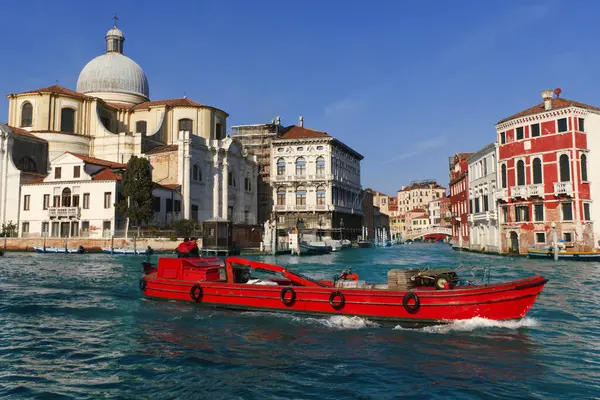 Canlı kırmızı bir tekne Venedik 'in turkuaz sularında geziniyor, tarihi binaların yanından geçiyor ve açık mavi gökyüzünün altındaki büyük kubbeli kilisenin önünden geçiyor..