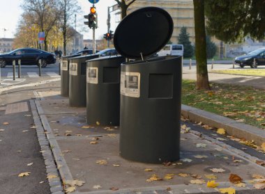 Kaldırımında açık kapağı olan modern, silindirik bir çöp kutusu. Sonbahar yaprakları etrafa saçılmış, geçen arabalar ve arka planda bir trafik ışığı..