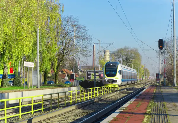 Modern bir tren bir istasyona açık bir günde yaklaşır, yemyeşil ağaçlarla çevrili, doğa ve kentsel altyapı arasındaki uyumu vurgular..