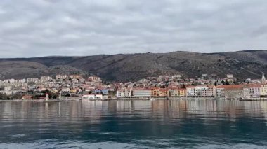 Hırvatistan 'ın Senj kentinin kıyı kasabasının panoramik manzarası, liman boyunca uzanan renkli binalar, bulutlu bir gökyüzünün altındaki engebeli tepeler tarafından destekleniyor ve sakin denize yansıyor..