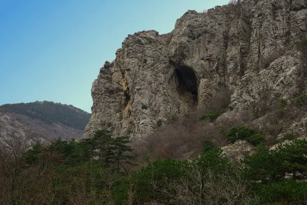 Görüntü, belirgin kayalık bir kayalık ve geniş bir mağaranın bulunduğu engebeli bir dağ manzarası yakalıyor, açık mavi gökyüzü altında yoğun bitki örtüsüyle çevrili, doğal güzellik ve macera sergiliyor..