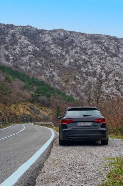 Pürüzsüz siyah bir araba kıvrımlı bir dağ yolunda, kayalık tepeler ve yeşilliklerle çevrili, parlak mavi bir gökyüzünün altında..