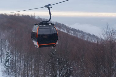 Modern bir teleferik kış ormanlarından yükseliyor, turuncu aksanları karlı araziye ve sisli dağ zeminine zıt düşüyor..