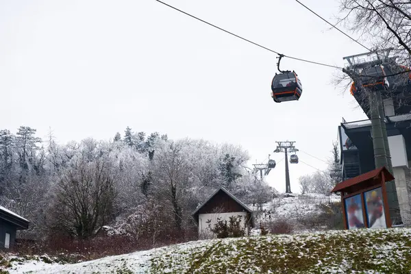 Yalnız bir teleferik karla kaplı bir ormanın içinden yükselir, bulutlu bir kış gününde, küçük bir binanın üzerinden geçer.