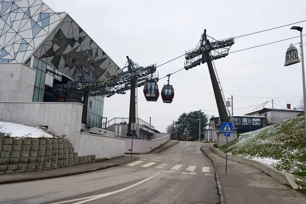 Geometrik cam mimarisi olan modern teleferik istasyonu, iki kırmızı gondol karlı, kentsel bir araziye giden bir yolun üzerinde asılı duruyor..