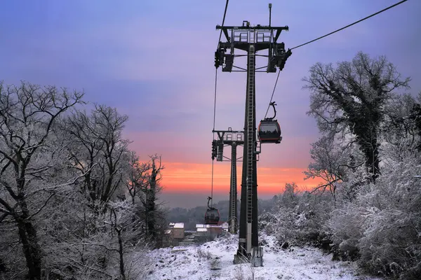 Bir teleferik, alacakaranlıkta karlı, ağaçlarla kaplı bir dağ manzarasına yükselir. Gün batımında canlı bir günbatımı, sıcak renklerle gökyüzünü boyar..