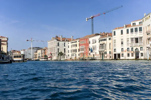 Venedik Büyük Kanalı manzaralı tarihi binalar rıhtımda, uzakta vinçler ve mavi sularda yavaşça yüzen tekneler..