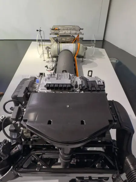 Bir platformda sergilenen modern bir araba motoru ve sürüş yağmurunun detaylı bir görüntüsü. Karmaşık bileşenler ve mühendislik, otomotiv iticilerinin arkasındaki teknolojiyi gözler önüne sererek vurgulandı..