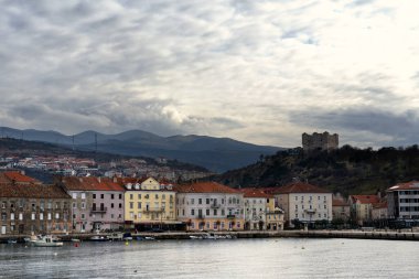 Senj, Croatia, showcases its historic charm with colorful buildings along the waterfront, a hilltop castle, and rolling hills under a dramatic, cloudy sky. clipart