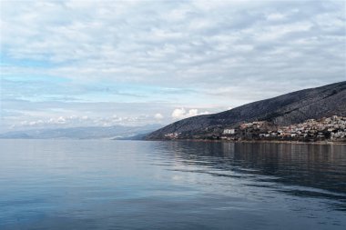 Hırvatistan 'ın Senj kenti, yumuşak ve dağınık bulutlarla dolu bir gökyüzünün altında, su kenarına uzanan bir yamaç kasabasıyla huzurlu bir kıyı manzarasını gözler önüne seriyor..