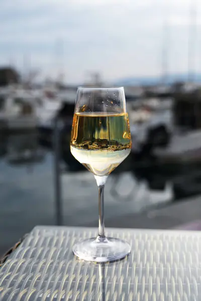 Bir bardak beyaz şarap, bulanık marina arkaplanlı dokulu bir masanın üzerinde duruyor. Bu da su kenarında rahatlatıcı bir an olduğunu gösteriyor..